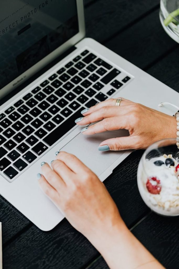 Close-up of womans hands on laptop and a bowl of yogurt with berries on a black table.